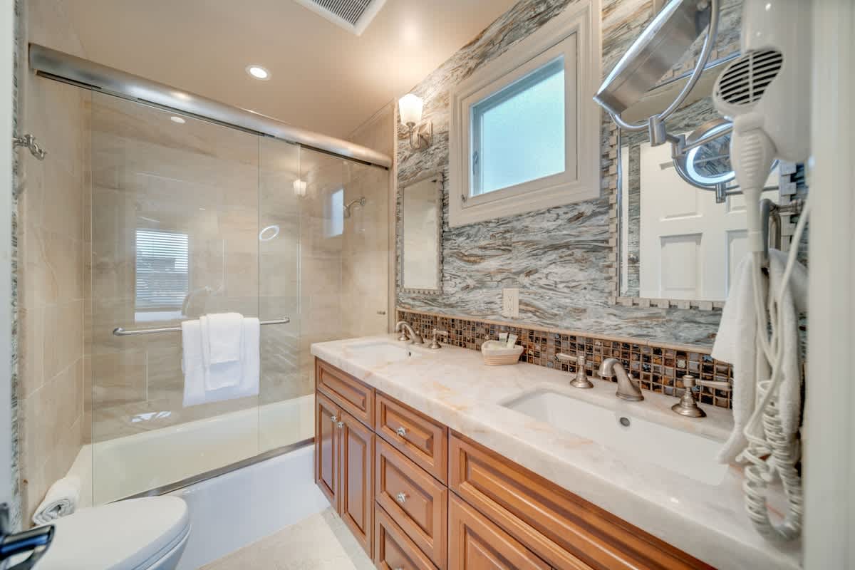 A modern bathroom with a double vanity featuring a light marble countertop, dark wood cabinets, and mosaic tile backsplash. It includes a shower-over-tub with a glass enclosure and a small frosted window.