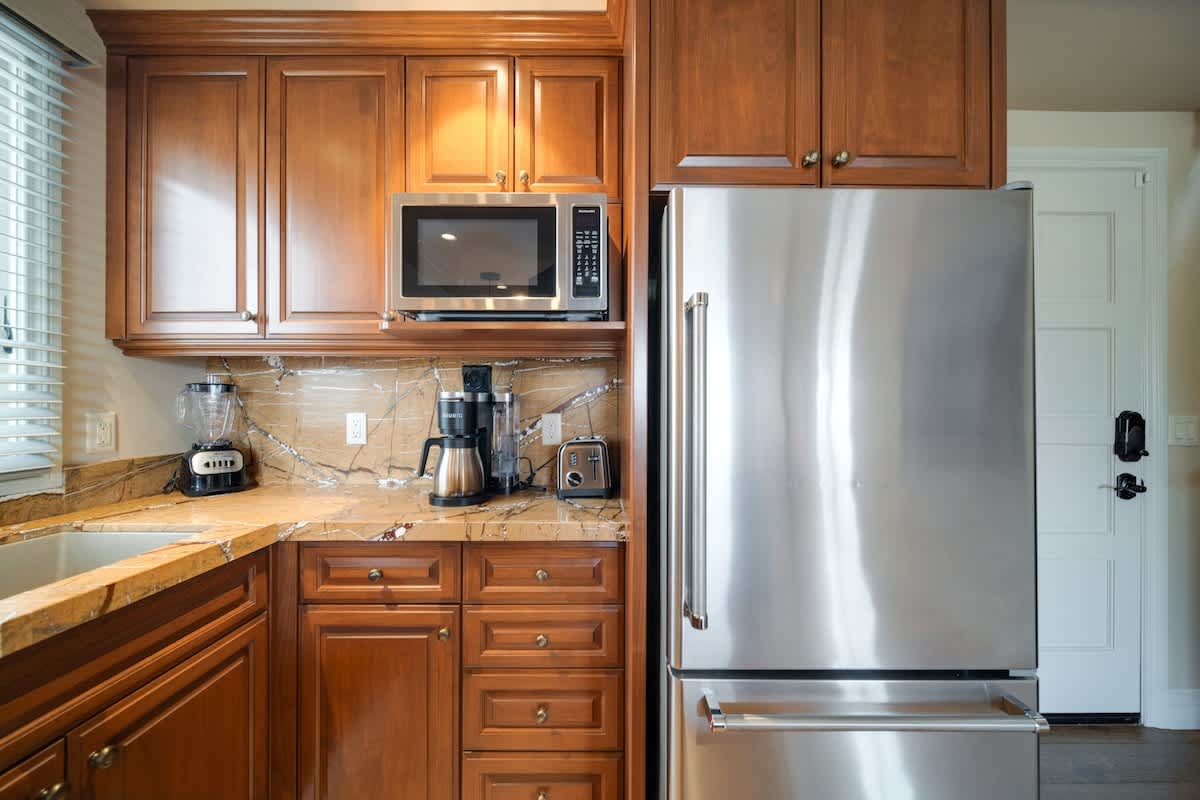 A well-equipped kitchen corner with rich cherry wood cabinets, granite countertops, and stainless steel appliances including a microwave, refrigerator, and coffee maker. A blender and toaster are also visible on the counter.