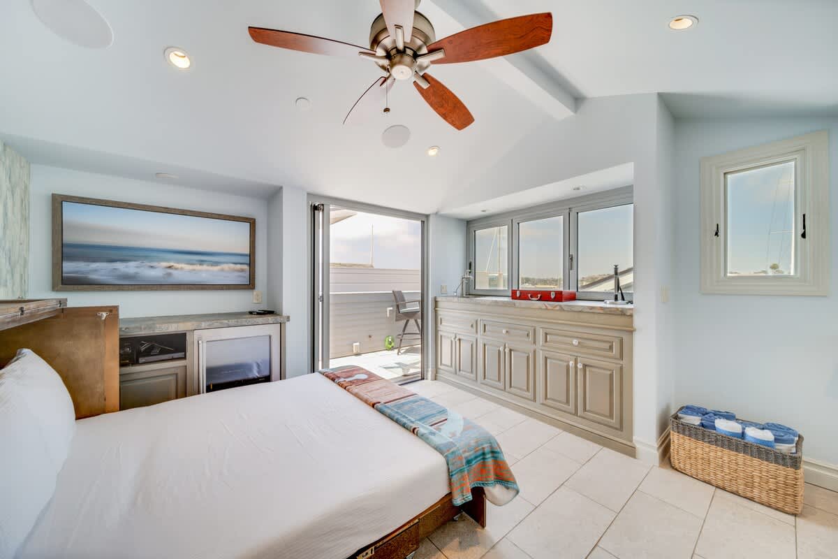Bright bedroom with a white bed, a large wall-mounted TV above a mini-fridge, and a built-in counter with windows, leading to a private balcony through sliding glass doors. A wooden ceiling fan hangs from the vaulted ceiling.