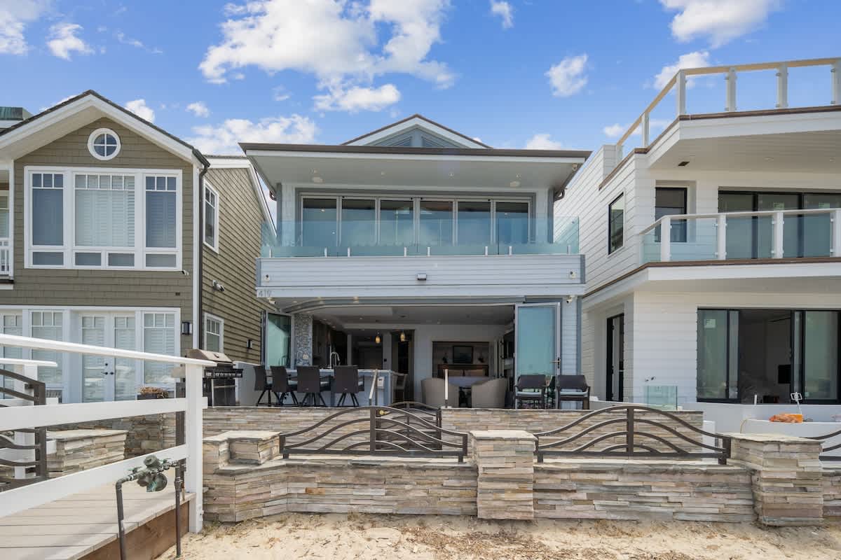 The modern back exterior of a multi-story house with large glass doors and balconies, overlooking a sandy area and the bay. A BBQ grill and outdoor seating are visible on the lower patio.