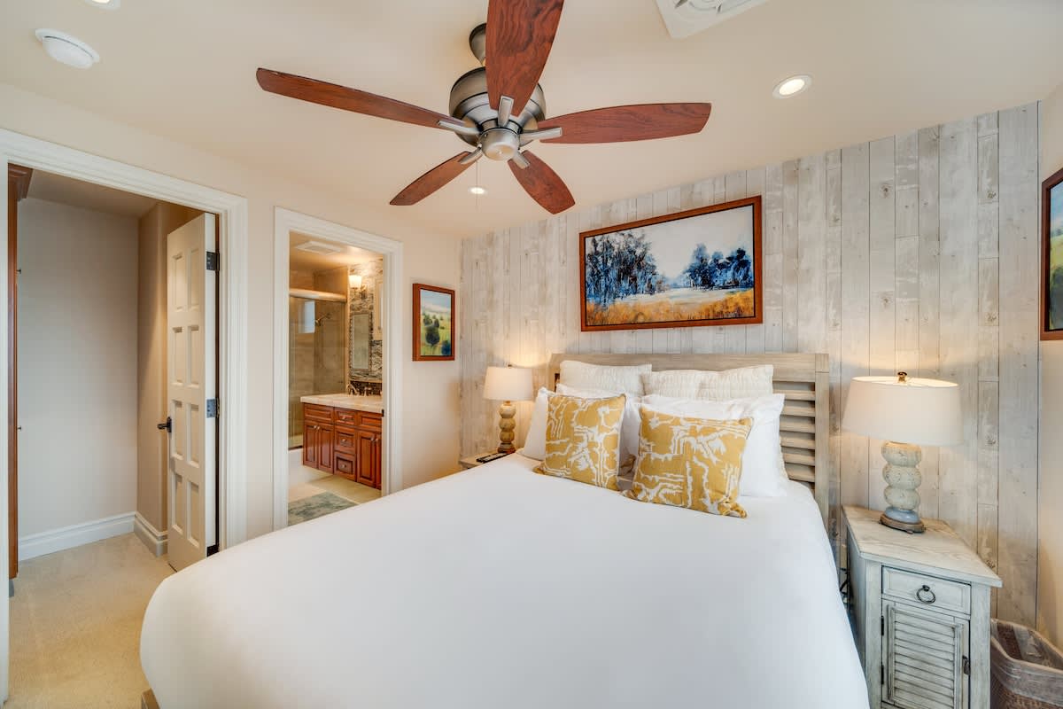 Another view of the bright bedroom with a queen bed, white bedding, and yellow pillows, showing a rustic nightstand with a lamp and an open doorway leading to a bathroom. The wall behind the bed features distressed white wood paneling.