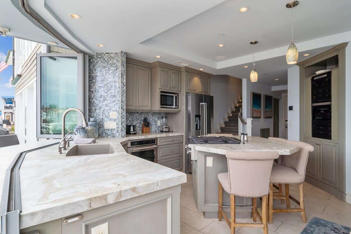 Modern kitchen featuring light gray shaker-style cabinets, white marble countertops, a mosaic tile backsplash, and stainless steel appliances, with a large island providing bar seating and a view towards a staircase.