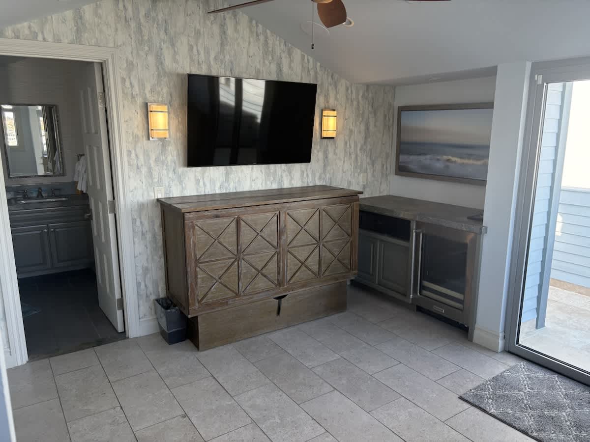 A versatile room with light tiled floors, a large wall-mounted TV flanked by sconces, and a wooden cabinet that may conceal a bed. The room also features a wet bar with a mini-fridge and an exterior door leading to an outdoor space.