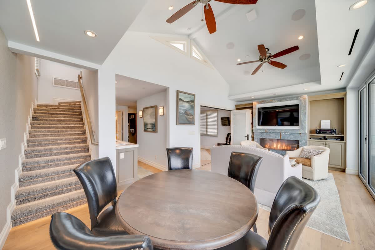 Spacious living area with light hardwood floors, a round dining table with black leather chairs, a modern fireplace with a TV above, and a carpeted staircase leading to an upper level, all under a vaulted ceiling.