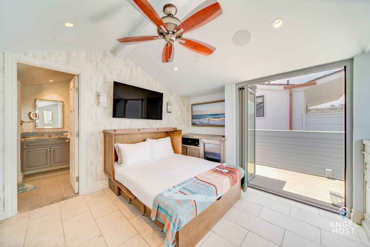 A versatile bedroom with tiled floors and a vaulted ceiling, featuring a murphy bed integrated into a wooden cabinet, a wall-mounted TV, and an en-suite bathroom visible through an open doorway, with access to an outdoor balcony.