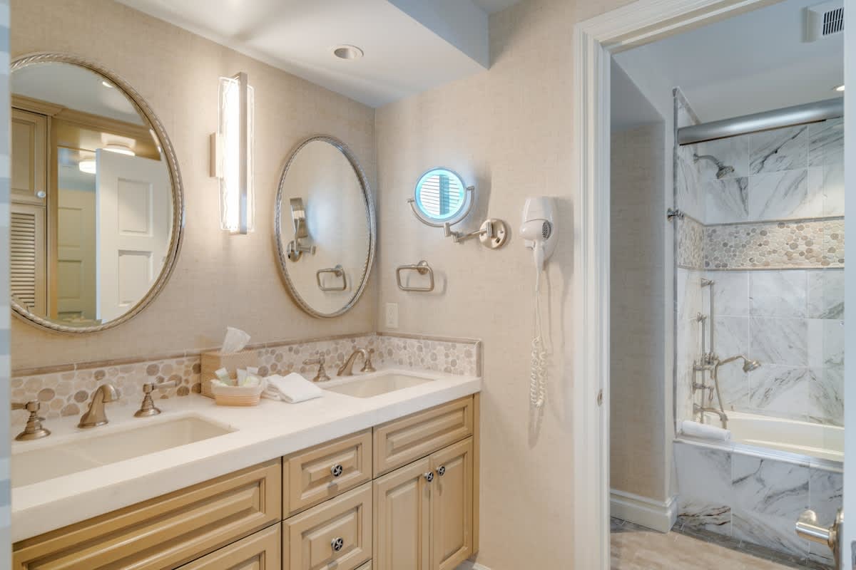 A spacious bathroom with a double vanity featuring light wood cabinets and a light-colored countertop with two oval sinks. Two oval mirrors with vertical sconces hang above, and a bathtub with a glass enclosure and marble-patterned tiles is visible to the right.