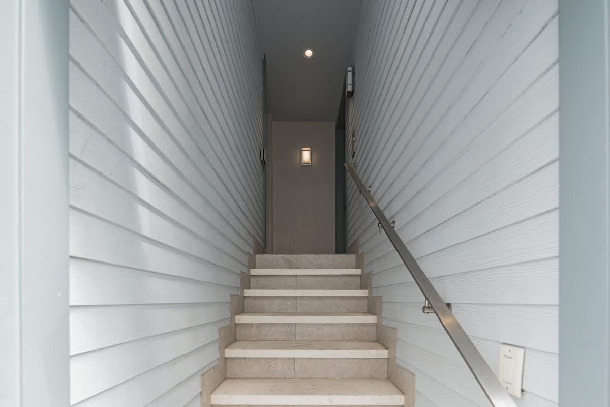A narrow, modern entryway with light-colored shiplap walls leading up a flight of stone stairs with a sleek metal handrail, illuminated by a small wall sconce at the top.