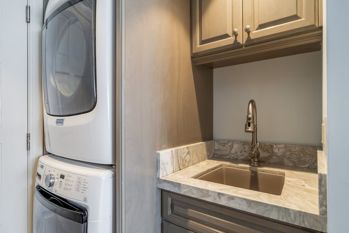A compact laundry area featuring a stacked white washer and dryer unit next to a small utility sink set into a light-colored marble countertop with a cabinet mounted above.