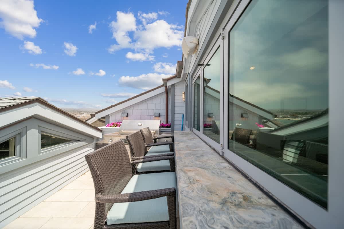A narrow balcony offering views of surrounding rooftops and distant water, furnished with several wicker chairs featuring light blue cushions lined up against a stone-topped railing. A built-in grill is visible at one end.