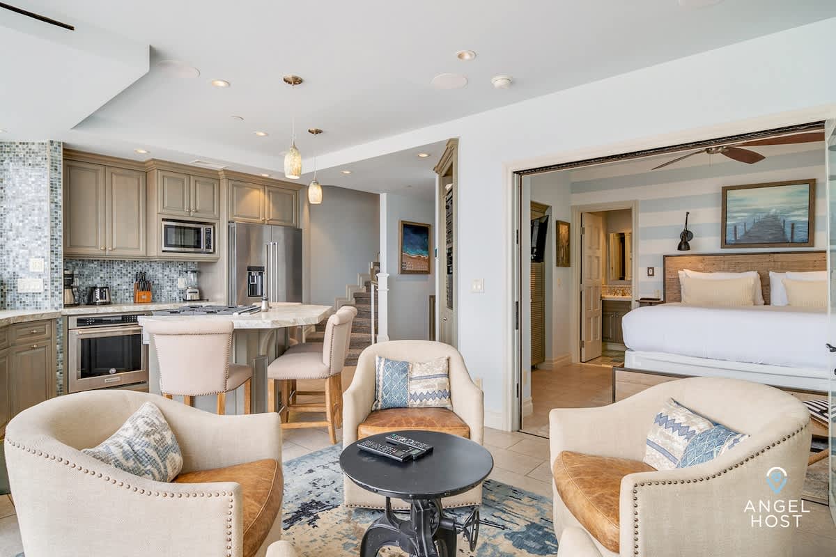 An open-concept living space with a seating area featuring three armchairs around a coffee table, flowing into a modern kitchen with light gray cabinets, stainless steel appliances, and a marble island with bar stools. A bedroom is visible through an open doorway.