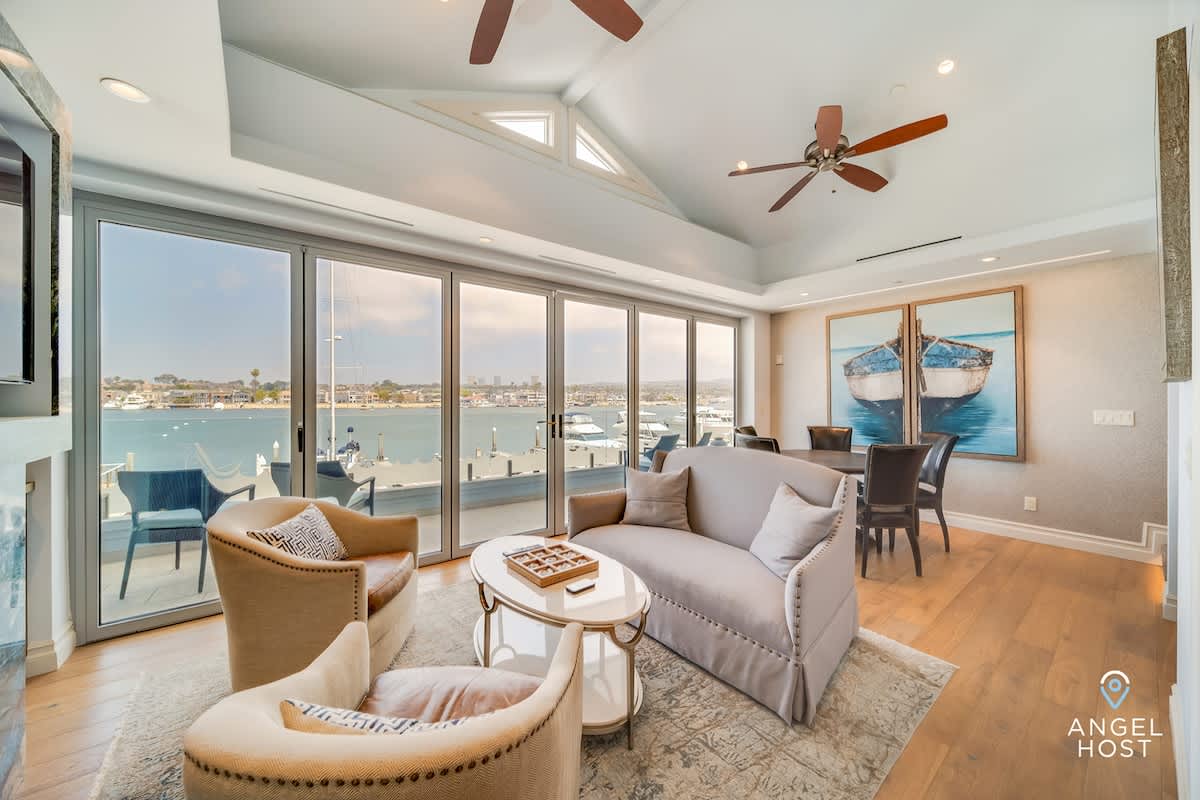 A spacious living room with hardwood floors and a vaulted ceiling, featuring a light gray sofa, two armchairs, and a coffee table, all facing large glass sliding doors that open to a waterfront view of boats and a city skyline.