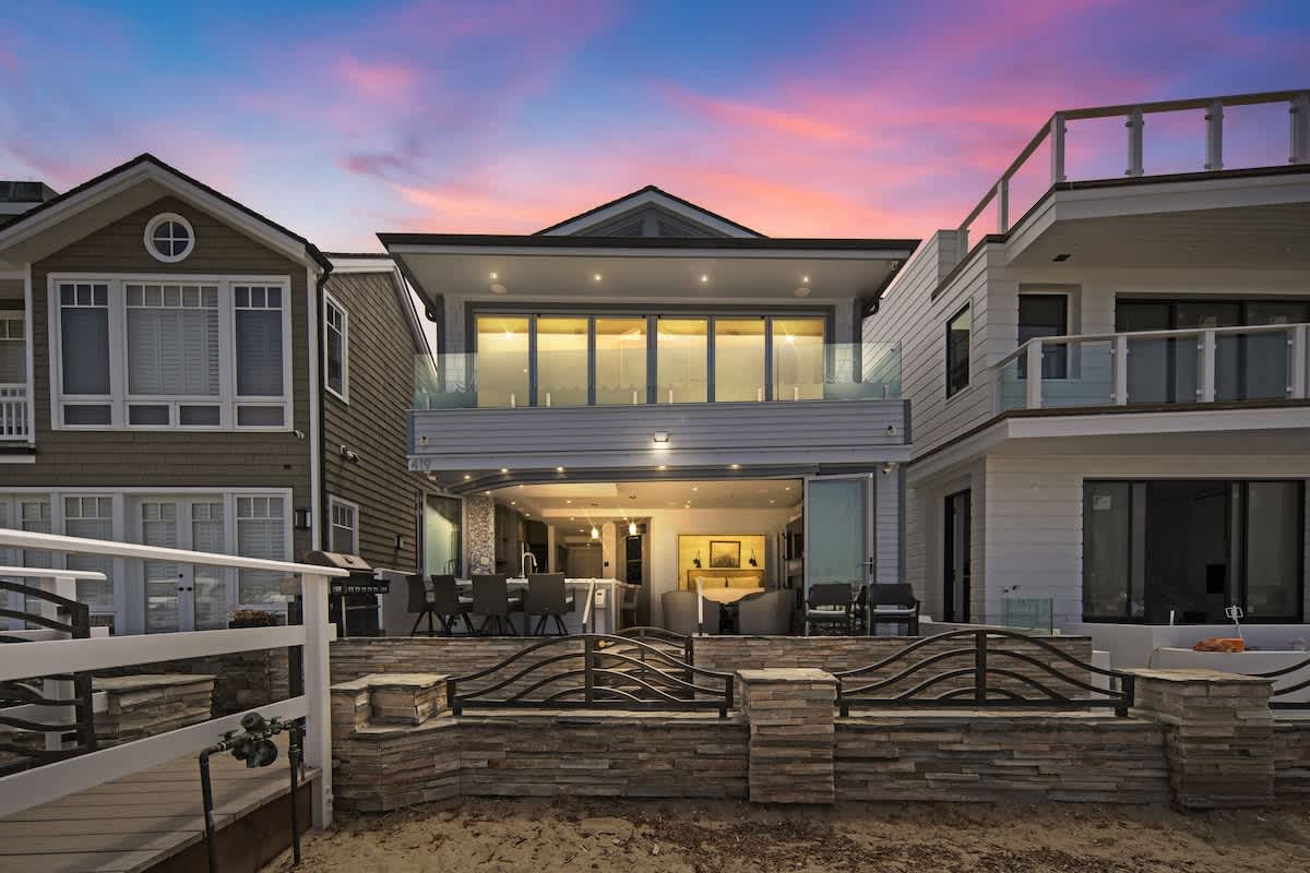 The back exterior of a modern house at dusk, featuring large glass sliding doors that open to a patio area with outdoor seating, overlooking a sandy beach and calm water. The upper level boasts a balcony with glass railings, all under a vibrant sunset sky.
