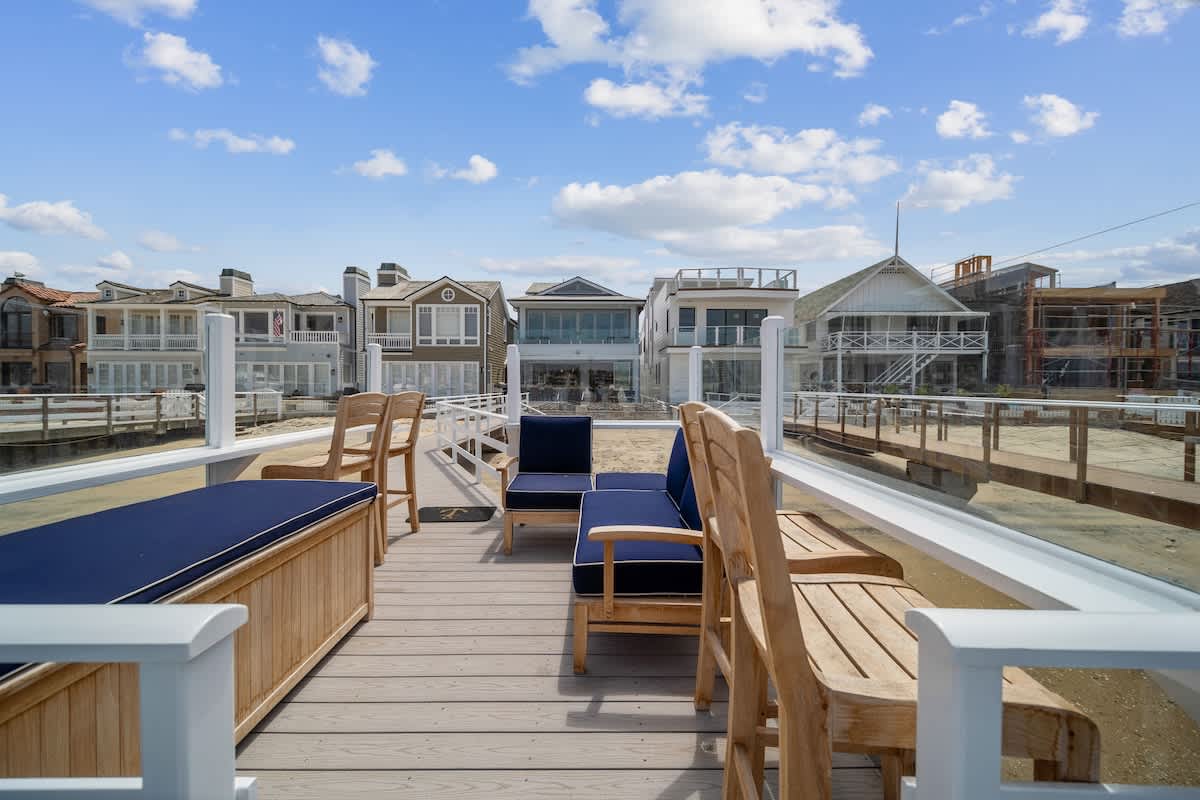 Spacious wooden deck with light wood outdoor chairs and benches featuring navy cushions, enclosed by glass railings, offering views of the bay, other waterfront homes, and boats docked nearby.