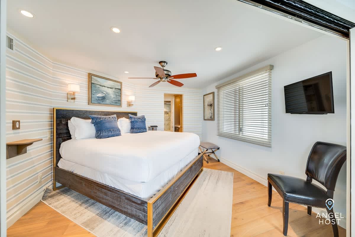 A cozy bedroom with light wood floors and a striped accent wall, featuring a large bed with a dark wood frame, white bedding, and blue throw pillows, along with a wall-mounted TV and a ceiling fan.