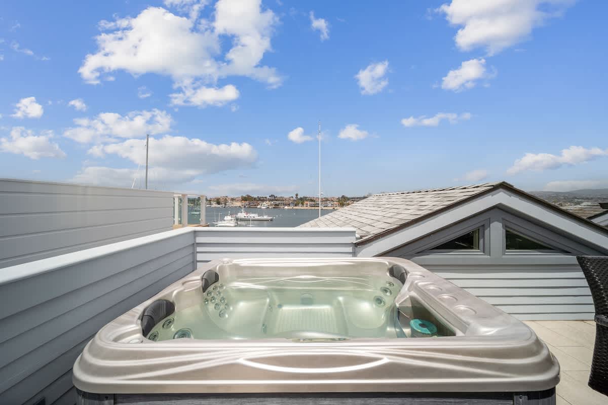 Outdoor hot tub situated on an upper deck with privacy walls, offering expansive views of the bay and distant cityscape under a bright blue sky with scattered clouds.