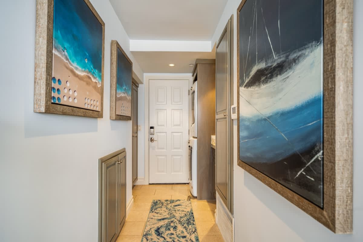 A bright hallway with light-colored walls and tiled floors, featuring two large framed artworks depicting beach scenes, and a discreet laundry area with a stacked washer and dryer near a white paneled door.
