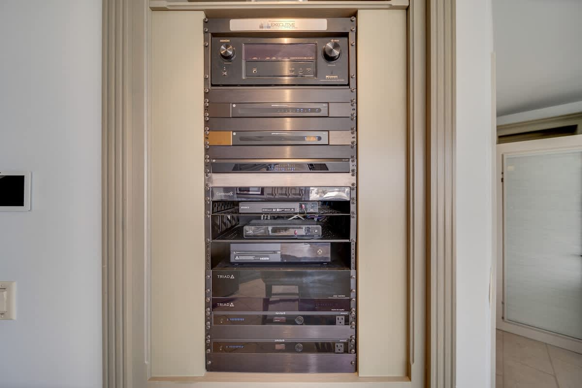 A tall, narrow built-in rack housing multiple black and silver electronic components, including an audio receiver, DVD players, and other media devices, set within a light-colored wooden frame.