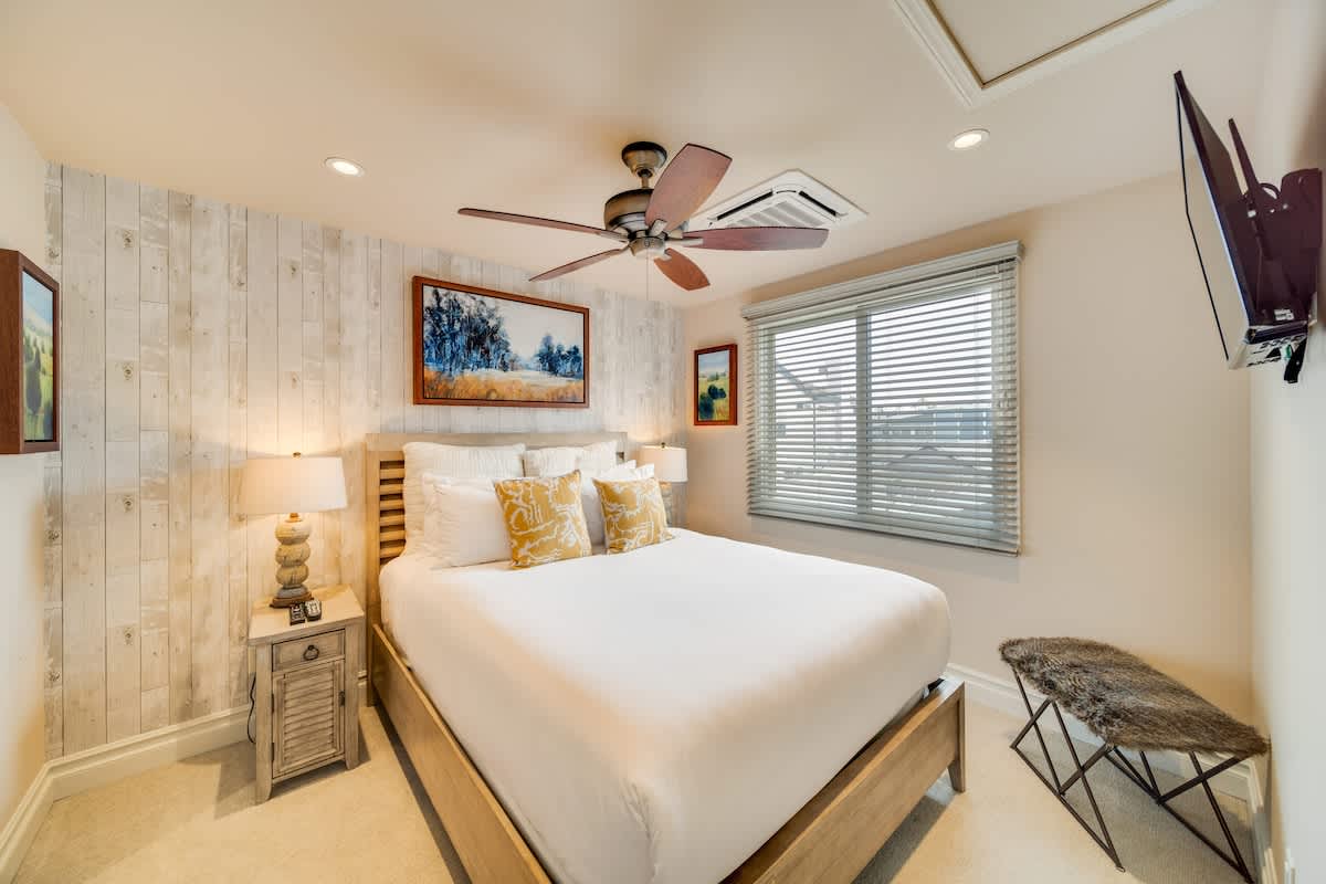 A bright bedroom featuring a queen-sized bed with crisp white bedding and yellow accent pillows, set against a light-toned shiplap wall. The room includes a rustic nightstand with a lamp, a ceiling fan, a window with blinds, and a wall-mounted television.