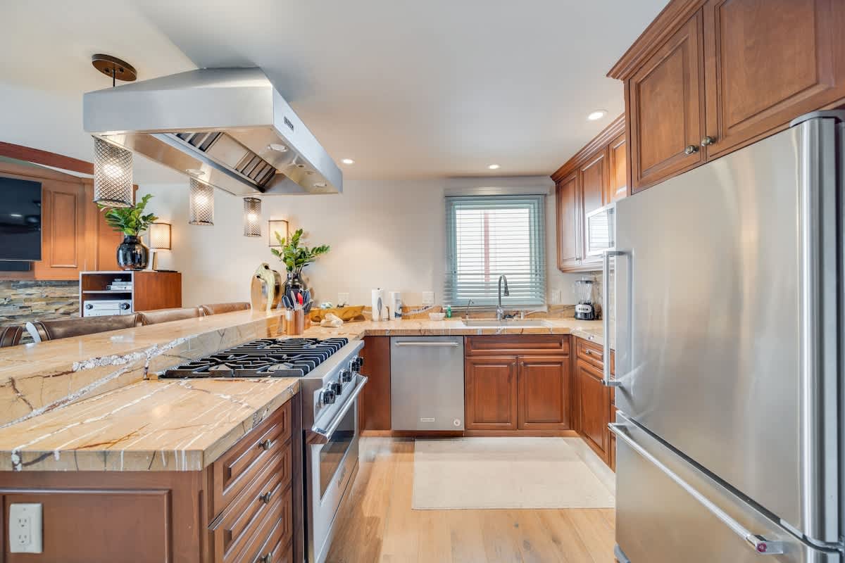Well-equipped kitchen with light wood cabinetry, granite countertops, a stainless steel gas range with a large hood, and a dishwasher. A large stainless steel refrigerator is visible on the right.