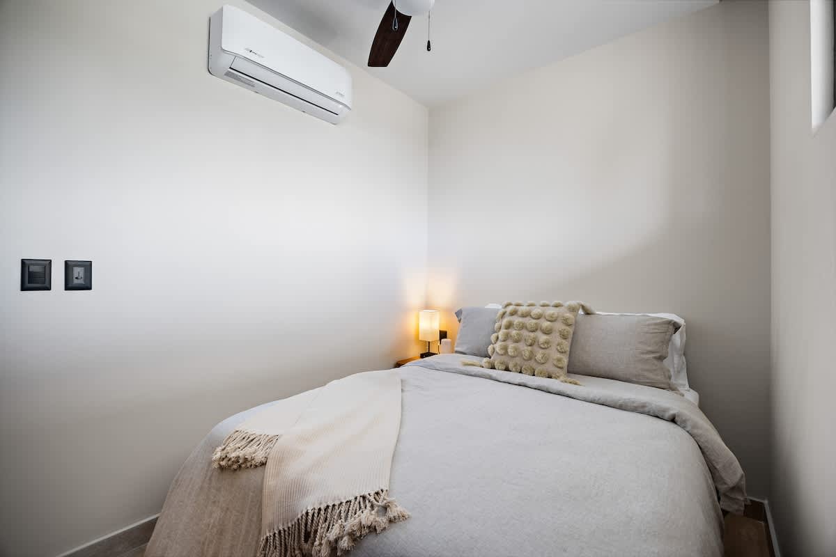 A cozy bedroom with light-colored walls, a queen-sized bed with light grey bedding, a cream-colored throw, and a decorative pillow. An air conditioning unit is mounted on the wall, and a ceiling fan hangs above.