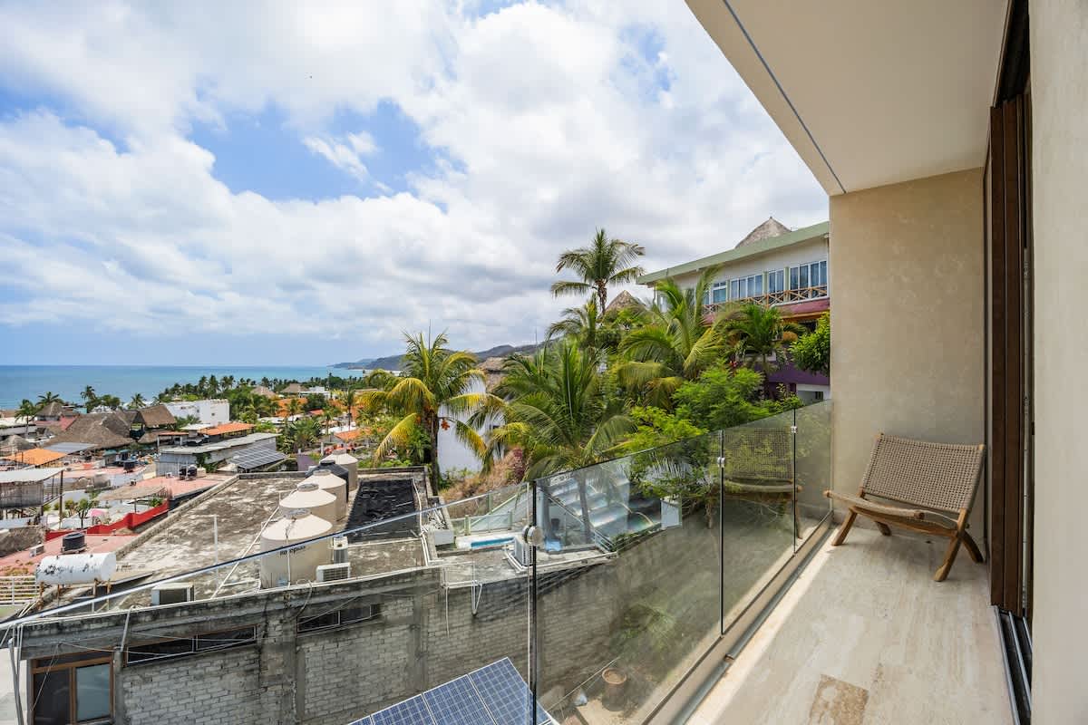 A serene balcony with a single woven chair offers a tranquil spot to enjoy the expansive views of the coastal town, lush palm trees, and the distant ocean under a bright sky. A glass railing ensures an open vista.