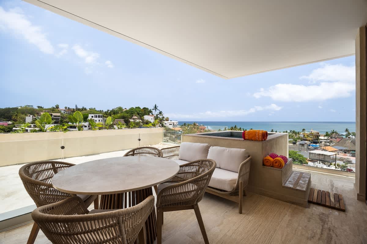 Spacious balcony featuring a round outdoor dining table with woven chairs and a built-in hot tub, offering stunning panoramic views of the ocean and the hillside.