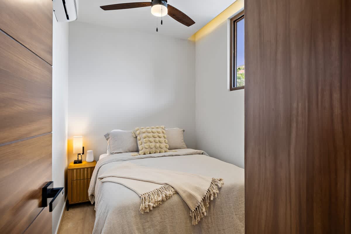 View into a compact bedroom with a queen bed, a wooden nightstand, and a lamp. A ceiling fan and an air conditioning unit are installed, and a window provides natural light.