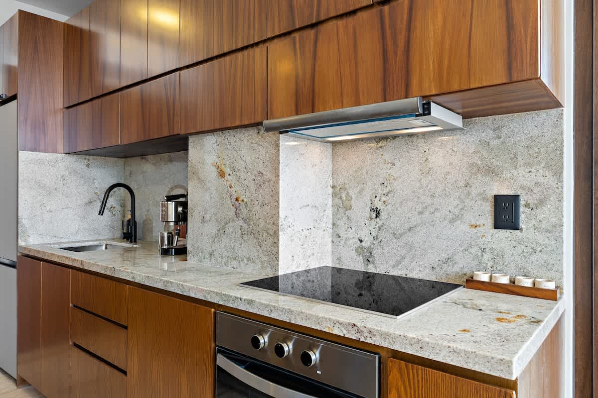 Modern kitchen with rich wooden upper and lower cabinets, light granite countertops and backsplash, a black induction cooktop, and a stainless steel range hood. A black gooseneck faucet and sink are visible on the left.