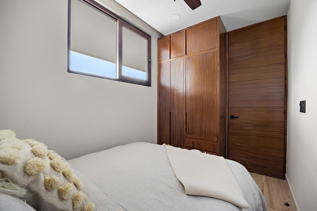A bedroom featuring a bed with light grey bedding, a window with blinds, and a large built-in wooden wardrobe and door, creating a warm contrast with the light walls.