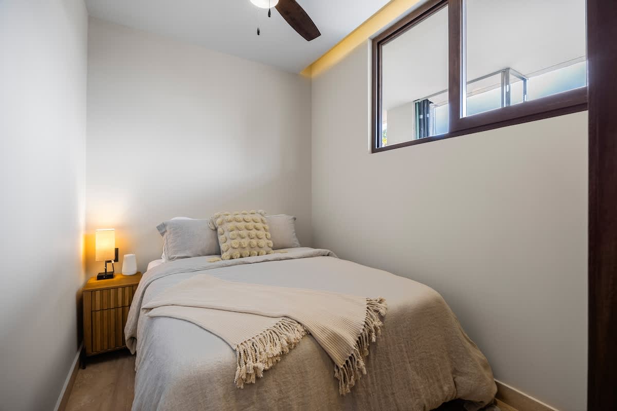 Cozy bedroom with a queen-sized bed dressed in light bedding, a wooden nightstand with a lamp, and a window on the right. A ceiling fan and an air conditioning unit are visible overhead.