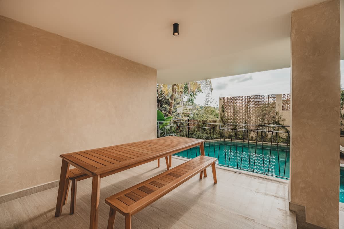A spacious covered patio features a long wooden picnic table with matching benches, positioned on light tiled flooring. Beyond a black metal railing, a swimming pool is visible, surrounded by lush tropical greenery and a partial view of the sky.
