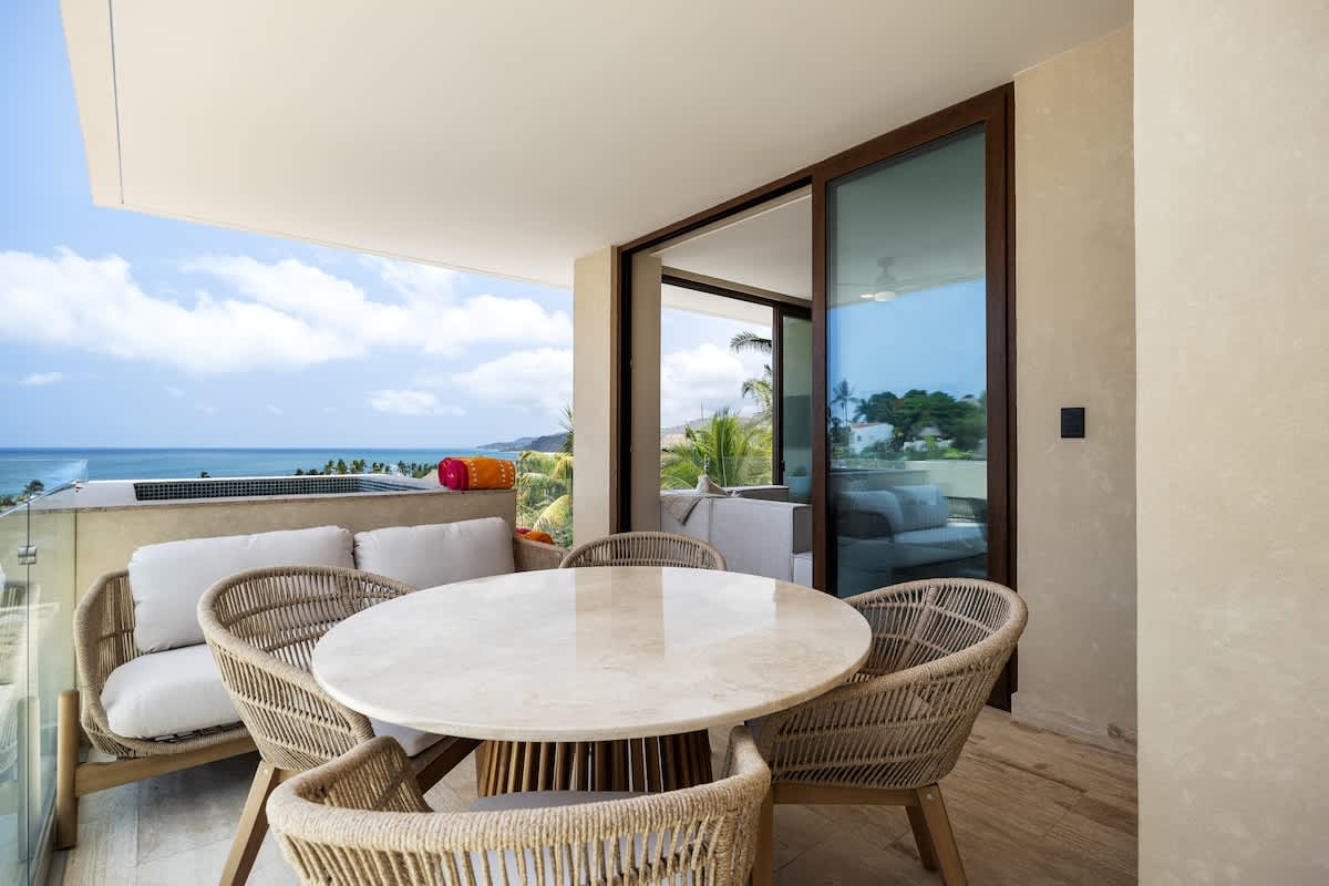 A spacious balcony features a round marble-top dining table with four woven chairs, a comfortable outdoor sofa, and a built-in hot tub overlooking the ocean. Large sliding glass doors connect to the interior.