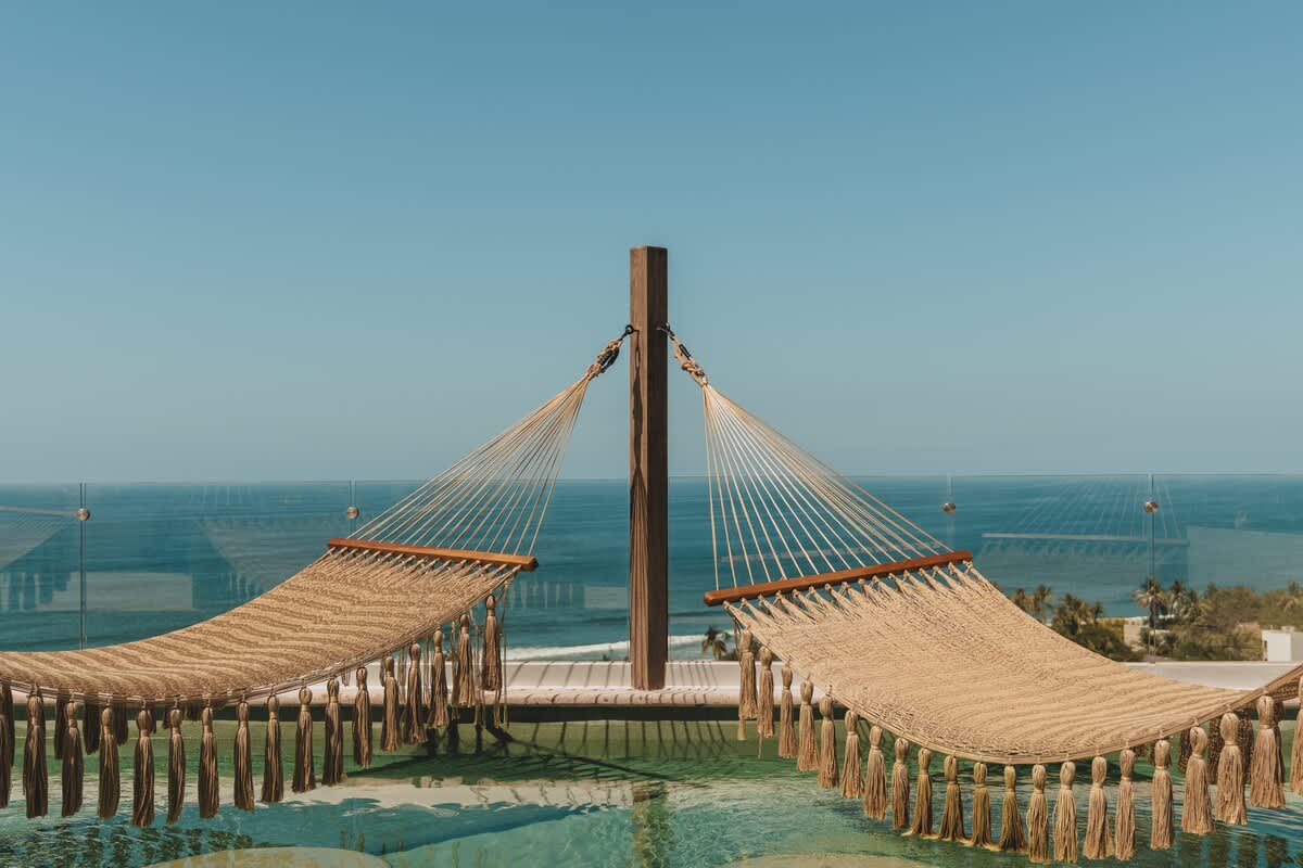 Two woven hammocks with tassels are suspended over a shallow pool, offering a unique lounging experience with a clear blue ocean and sky in the background.