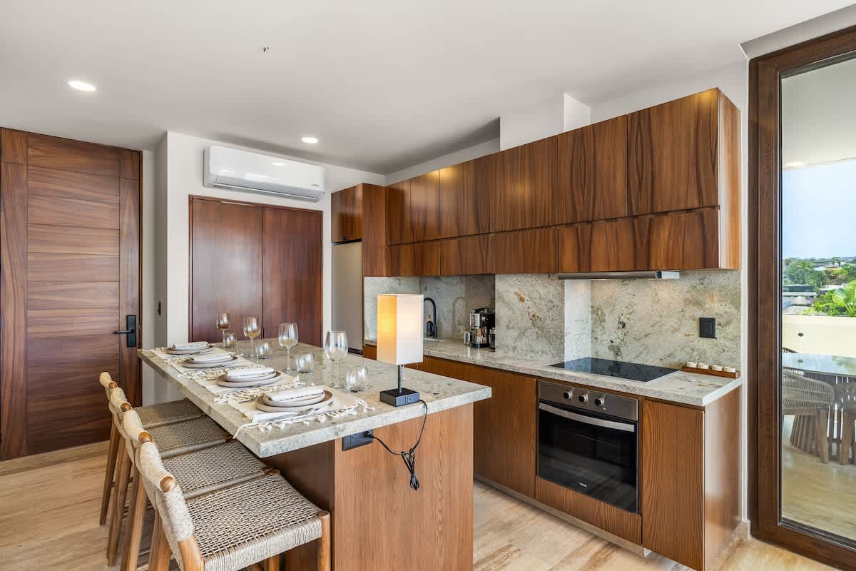 Wide shot of a modern kitchen featuring rich wooden cabinetry, light granite countertops, and a built-in oven. An island with a granite top serves as a dining area with four woven bar stools, set with plates and glasses.