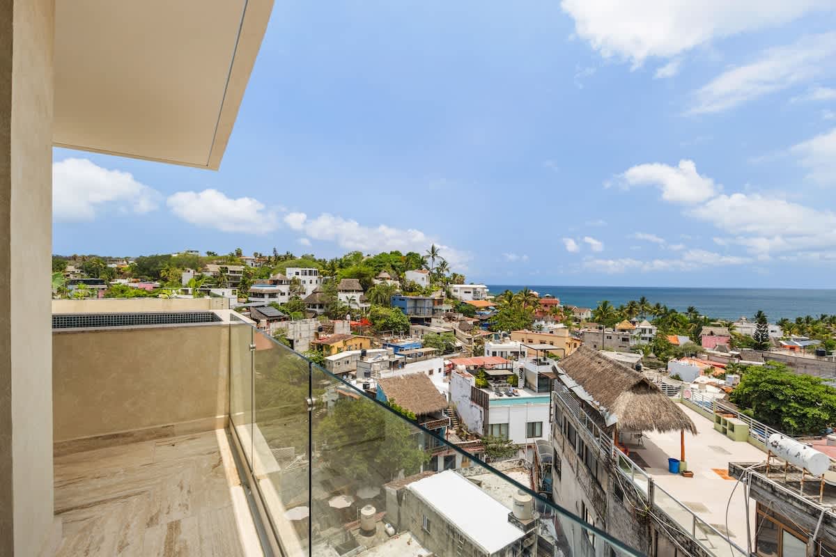A wide-angle view from a balcony showcases a sprawling coastal town with diverse rooftops and lush greenery, extending to the sparkling blue ocean under a partly cloudy sky. A clear glass railing provides an unobstructed panorama.