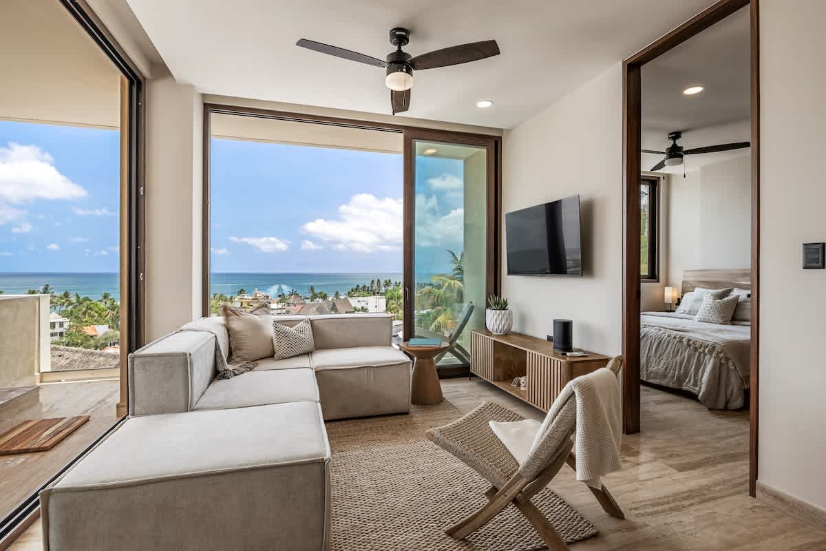 Contemporary living room with a light-colored sectional sofa, a ceiling fan, and large sliding glass doors opening to a balcony with ocean views. A flat-screen TV is mounted on the wall.