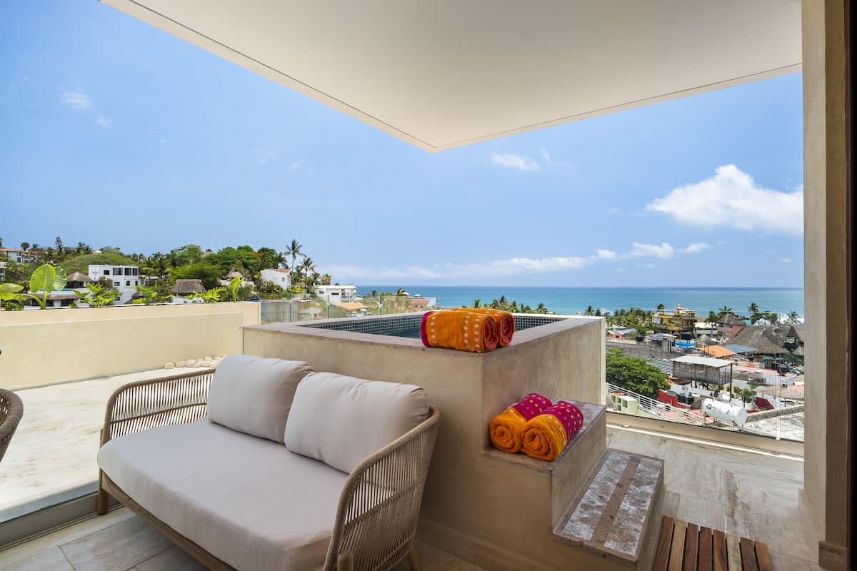 A comfortable outdoor sofa with light cushions sits on a balcony, adjacent to a built-in hot tub, both offering stunning panoramic views of the coastal town and the ocean horizon. Rolled towels are visible on the hot tub's edge.