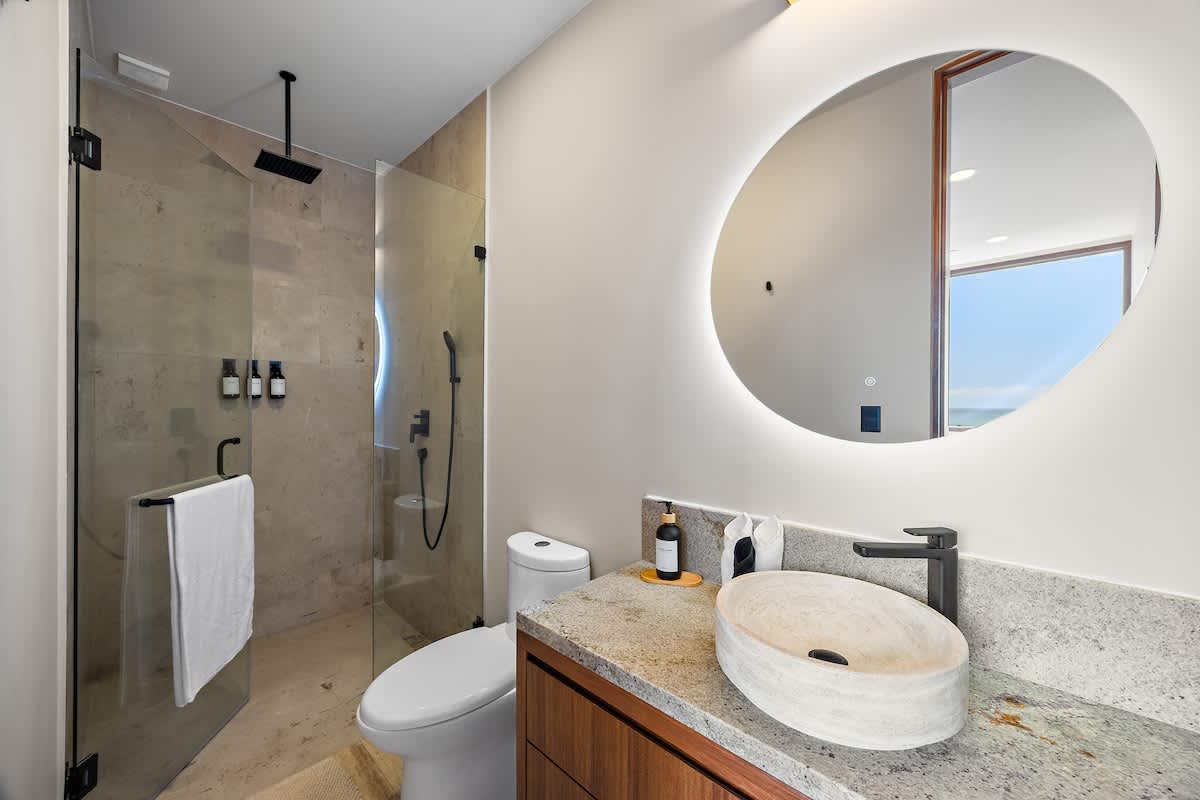 Modern bathroom featuring a walk-in shower with light beige tiles and black fixtures, alongside a vanity with a light granite countertop and a unique stone vessel sink. A large, round, backlit mirror hangs above the sink.