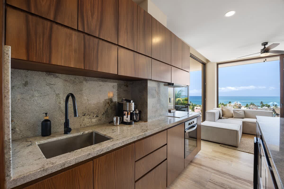 This contemporary kitchen features dark wooden cabinetry, light granite countertops, and a stainless steel sink with a black faucet, alongside a coffee machine. It offers an open view into the living area and out to the ocean.