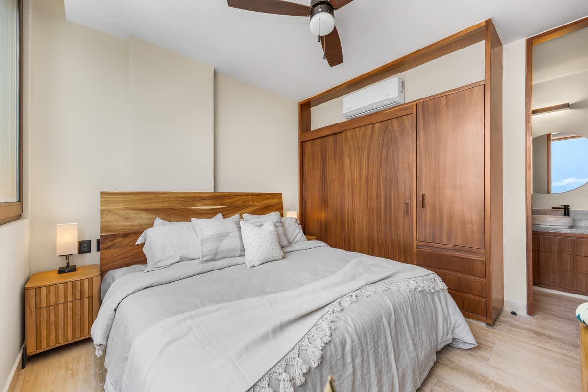 Spacious bedroom with a large bed featuring a wooden headboard and light grey bedding, flanked by wooden nightstands with lamps. A large wooden wardrobe and an air conditioning unit are built into the wall.