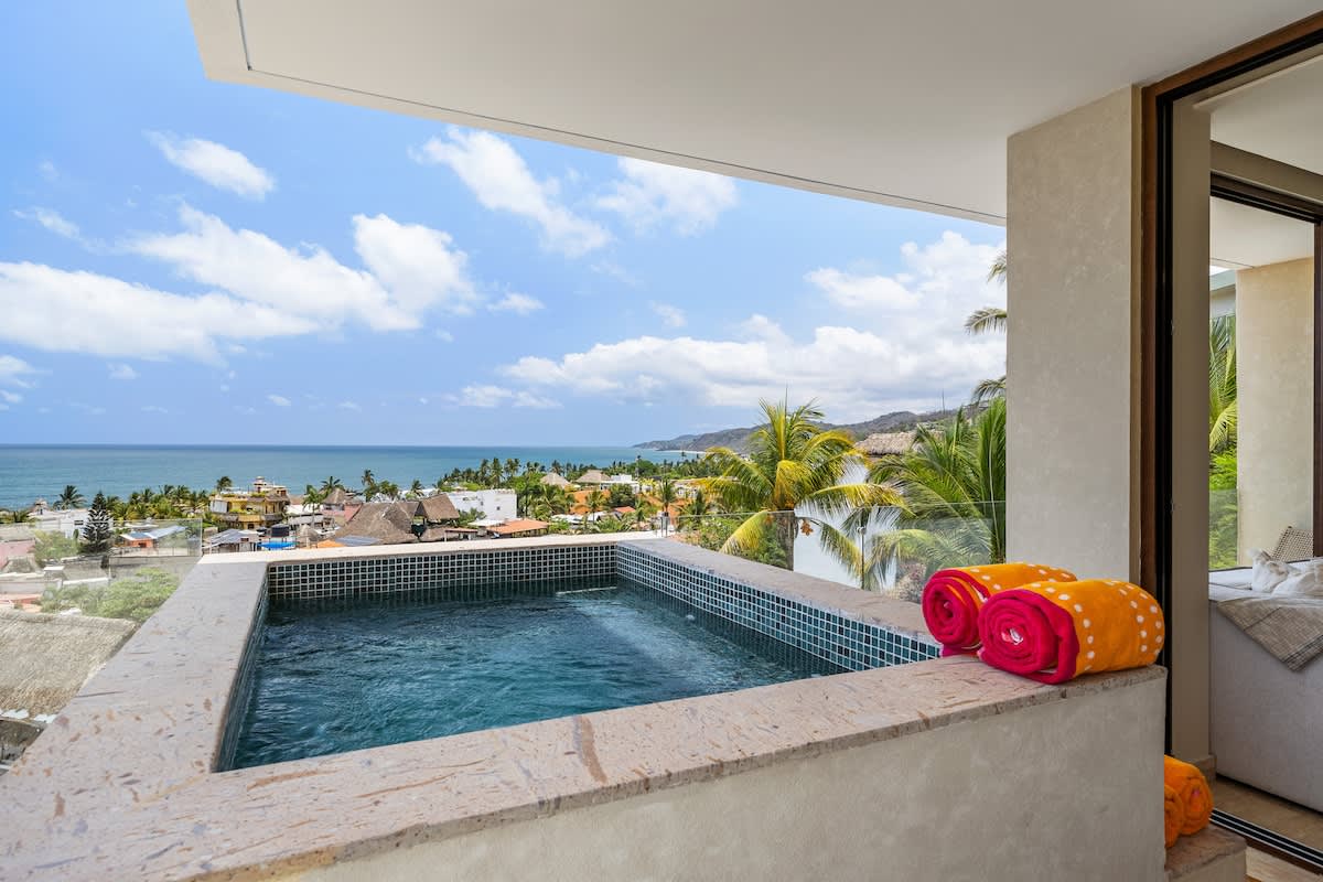 A built-in hot tub with blue mosaic tiles is prominently featured on a balcony, offering panoramic views of the ocean and a coastal town. Two orange and pink rolled towels rest on its stone edge.