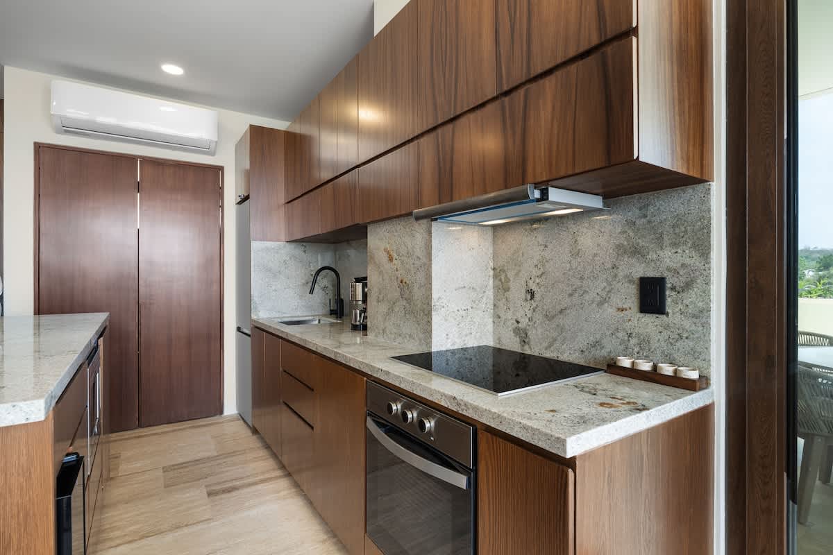 A sleek kitchen boasts rich wooden cabinetry and light granite countertops with a matching backsplash, featuring a built-in electric stovetop, oven, and a stainless steel sink with a modern black faucet. An air conditioning unit is mounted above.