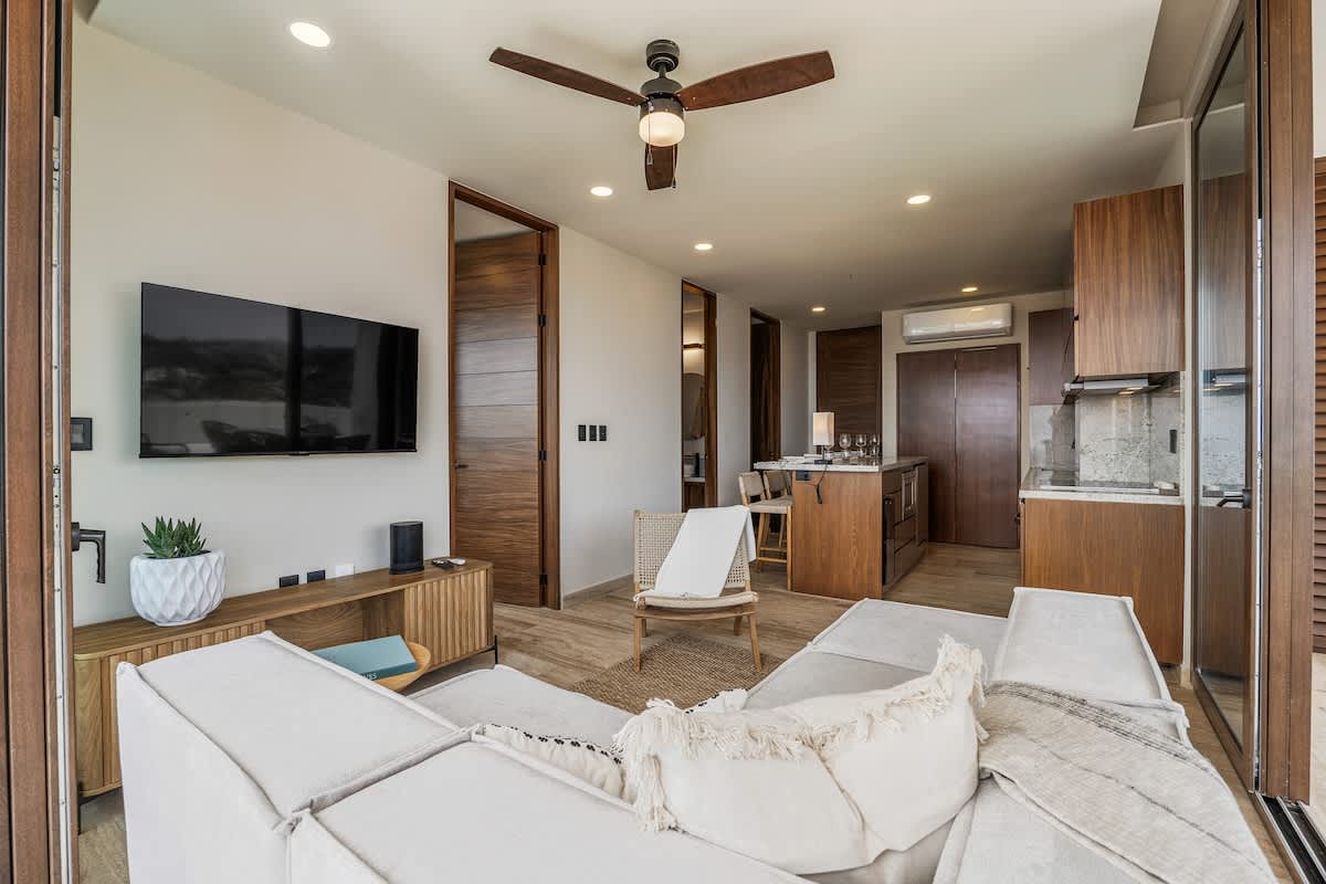 A modern living room features a large white sectional sofa facing a wall-mounted TV above a wooden console, complemented by a dark wood ceiling fan. Wooden doors and a partial view of the kitchen island are visible in the background.