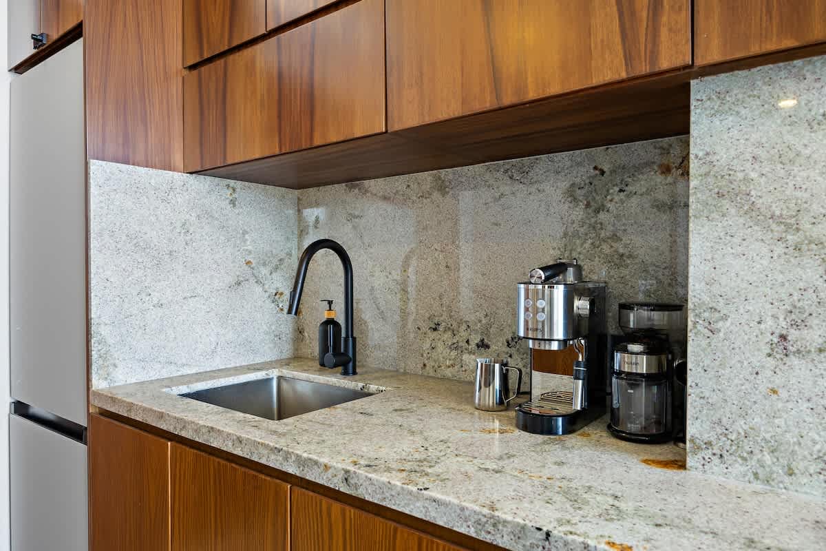 Close-up of a kitchen corner with a stainless steel sink and a black gooseneck faucet, set against a light granite countertop and backsplash. A modern coffee maker and grinder are placed next to the sink.