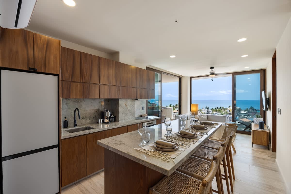 Modern kitchen with dark wood cabinets, light stone countertops, and a large island serving as a dining space, seamlessly connecting to the living area with ocean views.