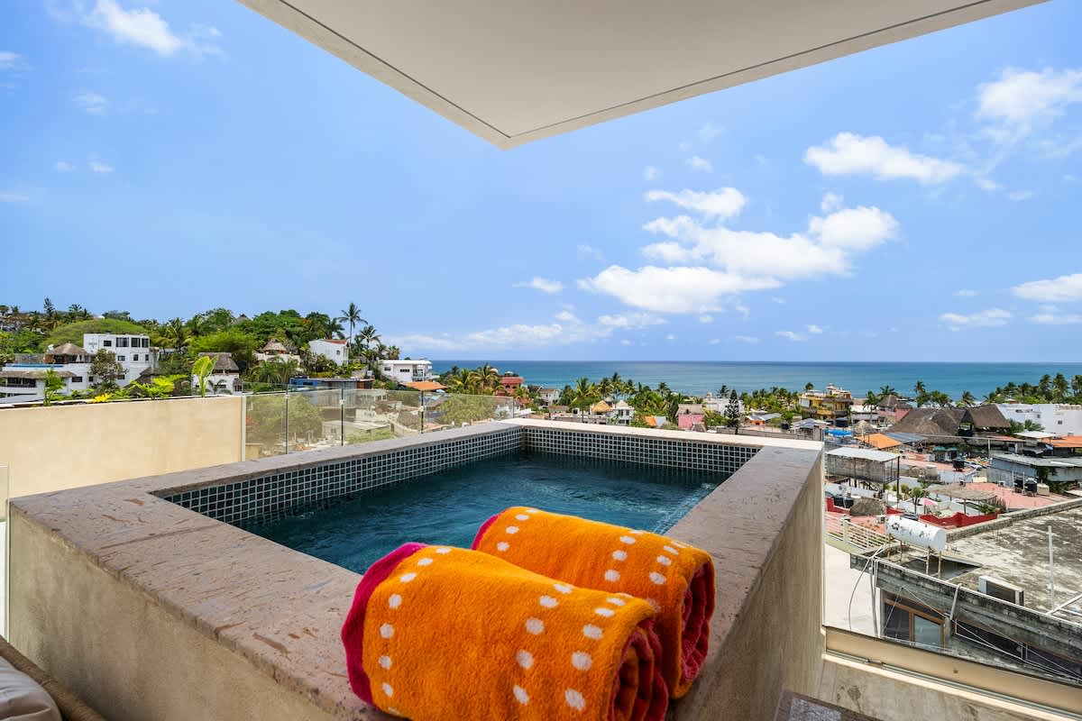 This elevated hot tub on a balcony provides expansive views of the vibrant coastal town below and the vast blue ocean beyond. Two colorful rolled towels are placed on the hot tub's edge.