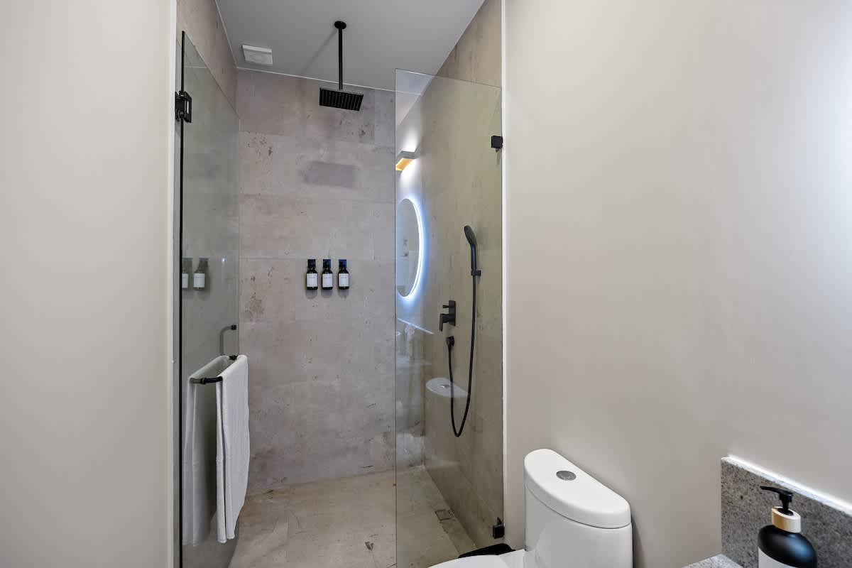 Modern bathroom featuring a walk-in shower with light tiled walls and black fixtures, including a rainfall showerhead. A toilet is visible next to the glass shower enclosure, and a backlit mirror is reflected in the glass.