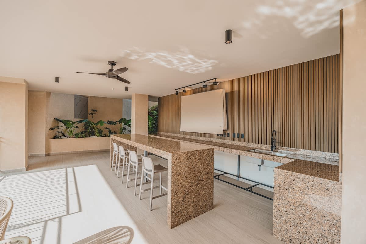 A spacious covered outdoor area features a long, speckled granite bar with multiple bar stools, complemented by a slatted wooden accent wall and a projector screen. Sunlight streams onto the light tiled floor, and lush plants are visible in a planter to the left.