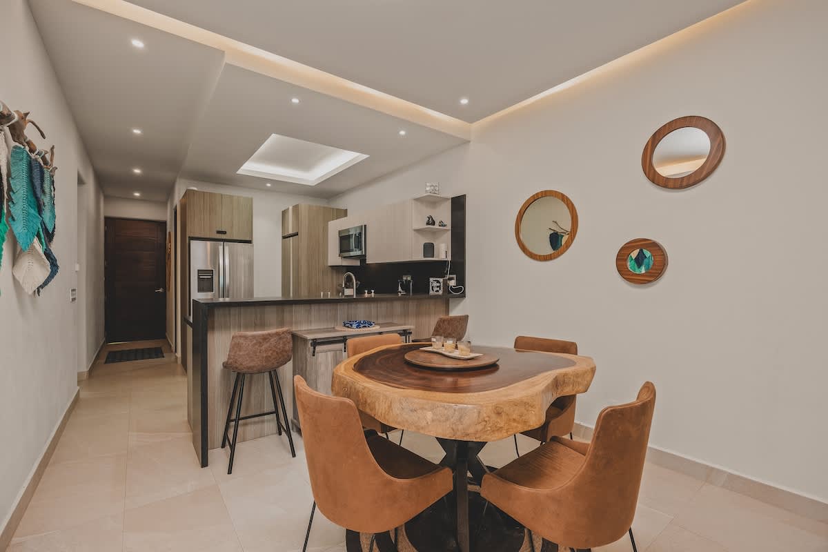 View from the dining area towards the kitchen, showing a unique live-edge wood dining table with brown leather chairs. The modern kitchen features light wood cabinetry, dark countertops, and a breakfast bar.