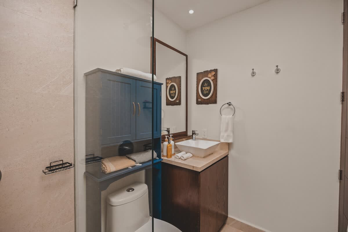 View of a contemporary bathroom with a dark wood vanity, white vessel sink, and a blue storage cabinet above the toilet, separated from the shower by a glass panel. Decorative wall art and white towels add a homely touch.